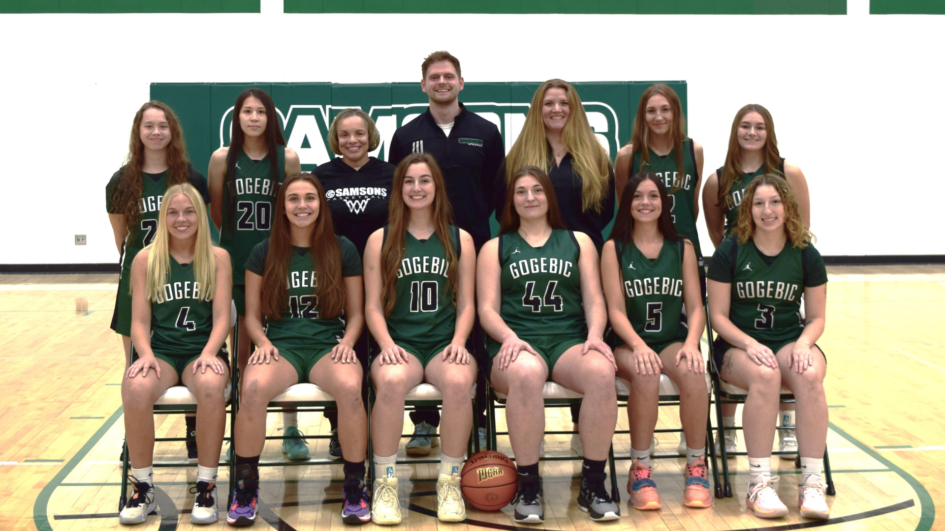 Women basketball players sitting in chairs and standing, in a team photo with a basketball at their feet.